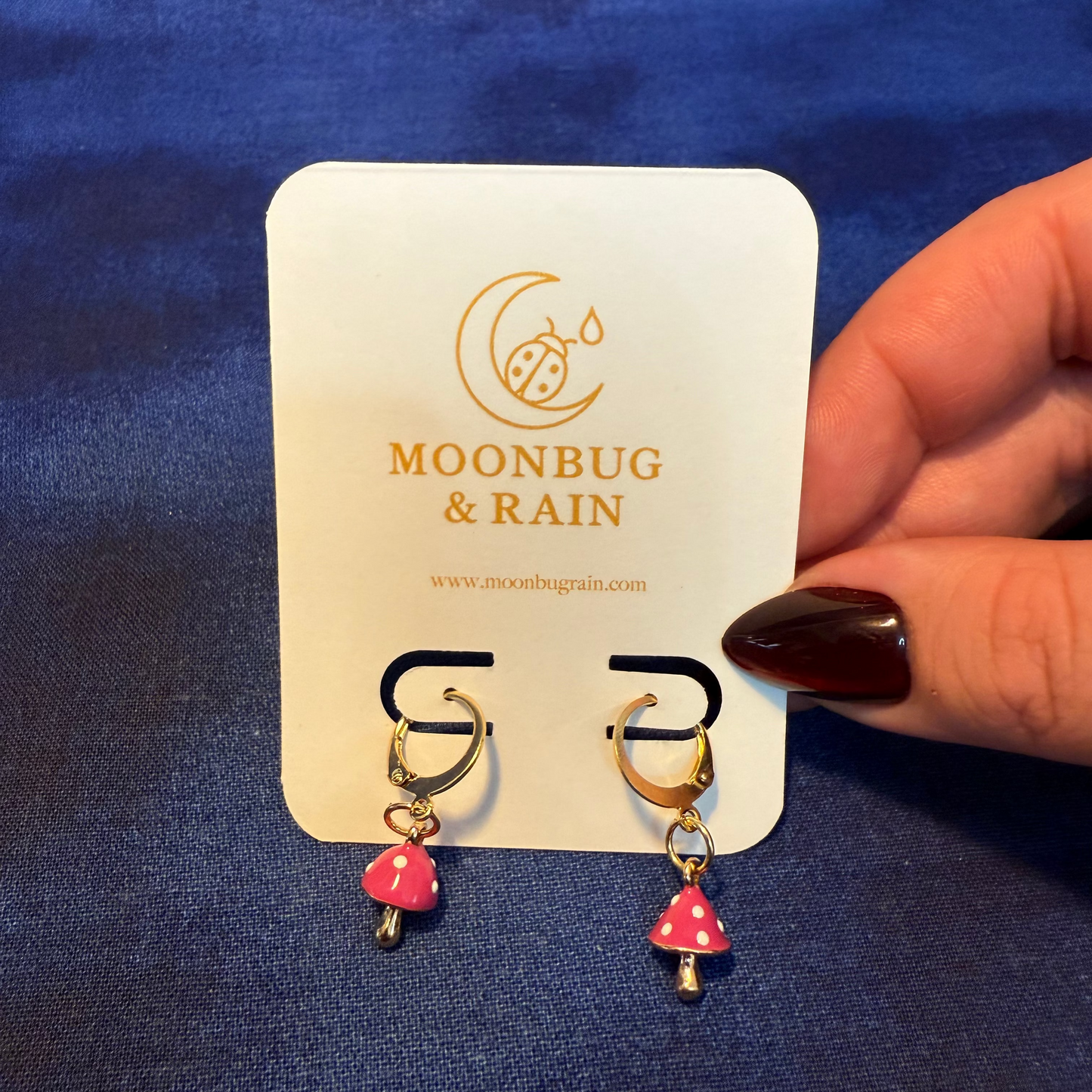 Pair of mushroom-shaped earrings on a 'Moonbug & Rain' branded card against a blue background.