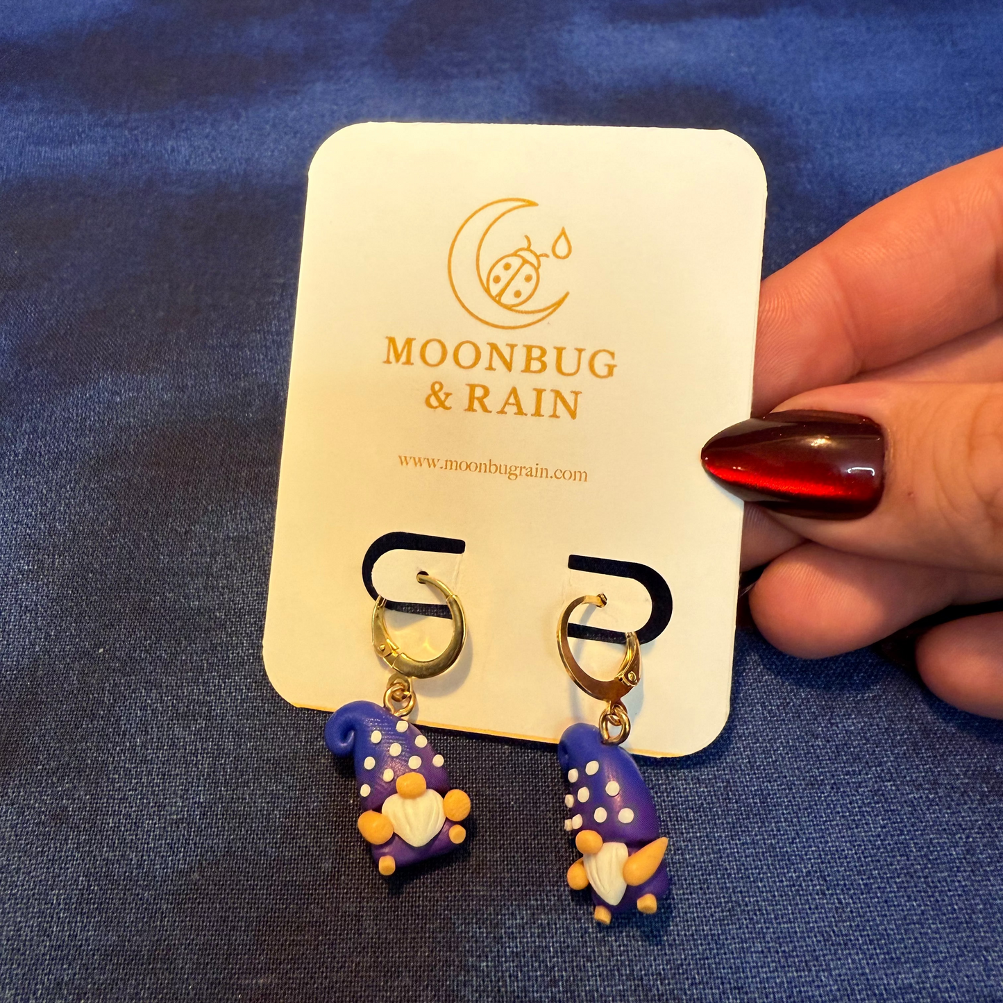Pair of blue and gold earrings on a 'Moonbug & Rain' branded card held by a hand with red nail polish.