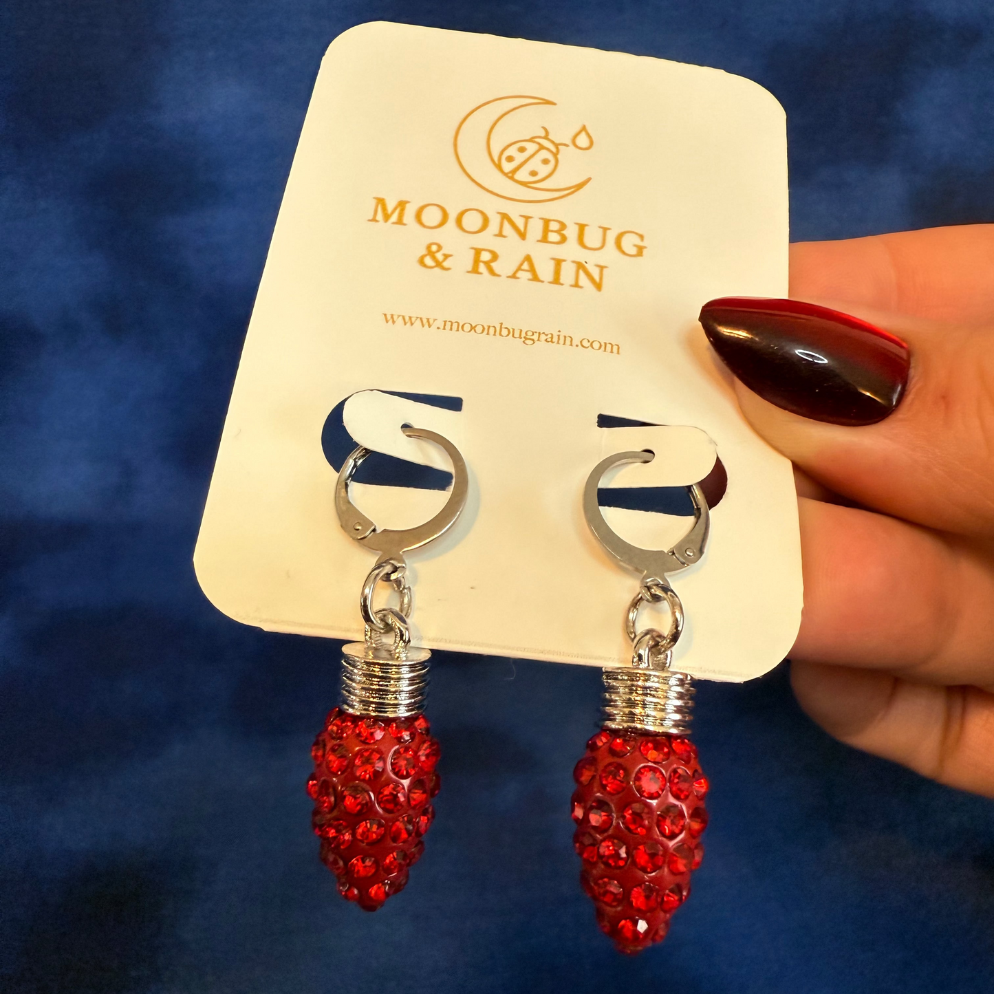 Red earrings with silver accents on a 'Moonbug & Rain' branded card against a blue background.