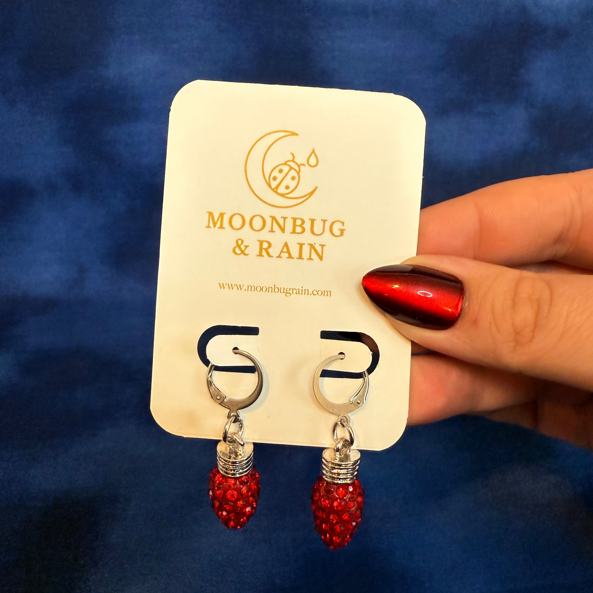 Red earrings on a 'Moonbug & Rain' branded card against a blue background