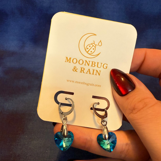 Heart-shaped earrings with blue gemstones on a 'Moonbug & Rain' branded card.