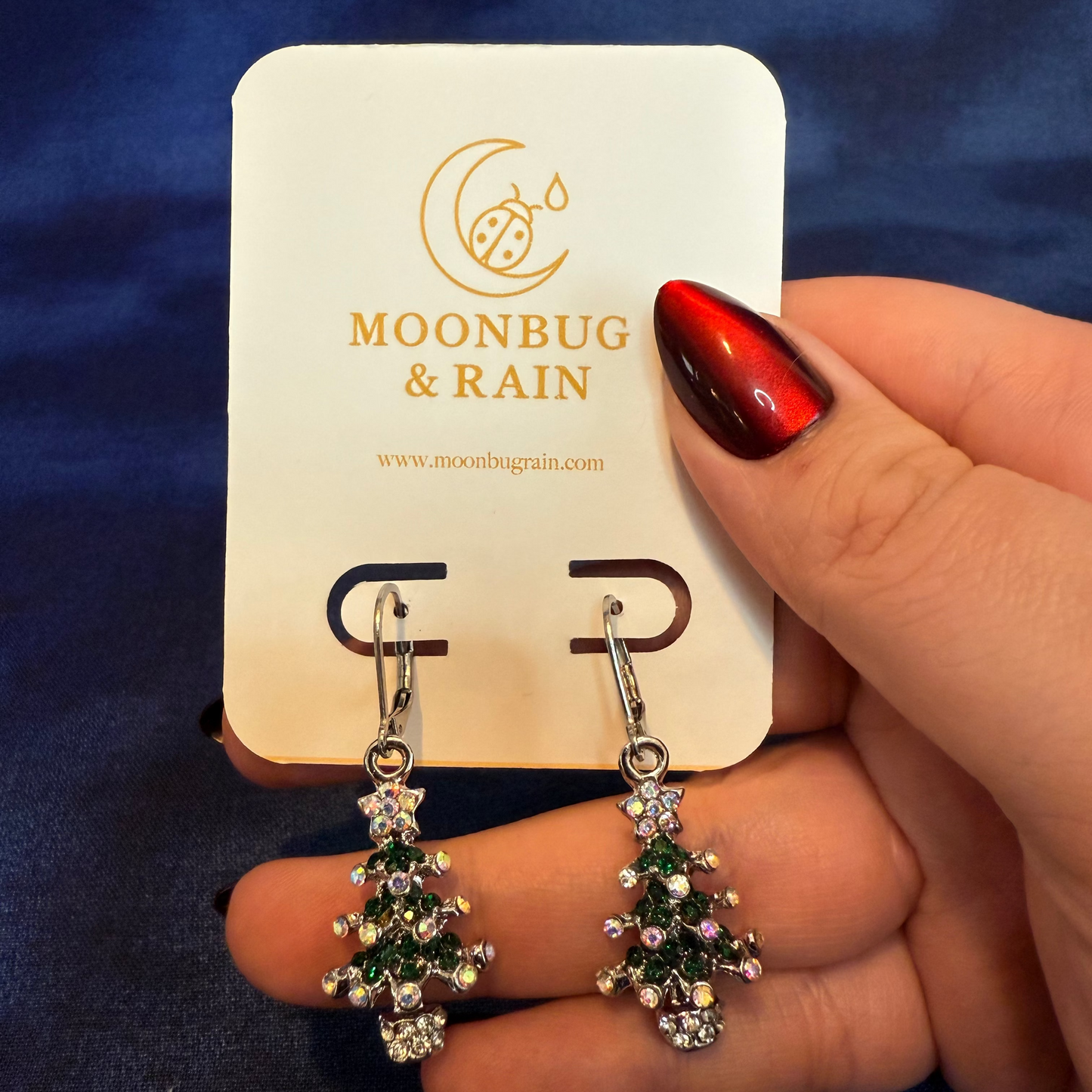 Tree-shaped earrings held by a hand on a Moonbug & Rain card against a blue background