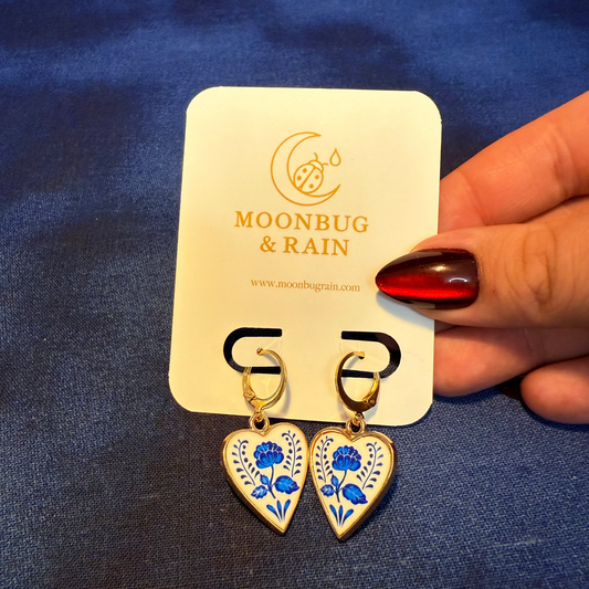 Heart-shaped earrings with blue floral designs on a 'Moonbug & Rain' card against a blue fabric background.