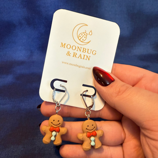Gingerbread man earrings held by a hand with a 'Moonbug & Rain' card in the background.