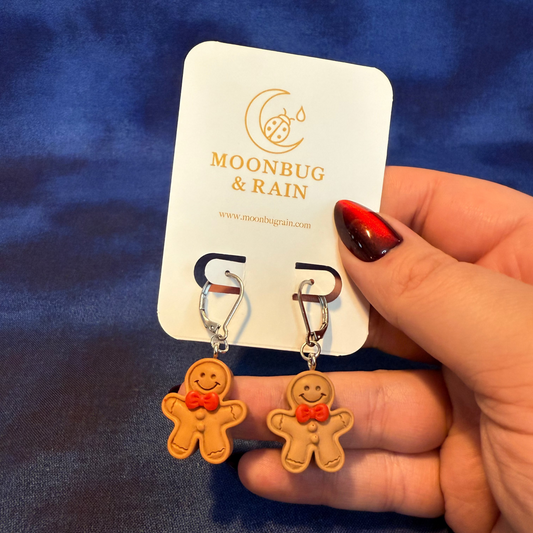 Gingerbread man earrings held by a hand with red nail polish against a blue background, branded 'Moonbug & Rain'.