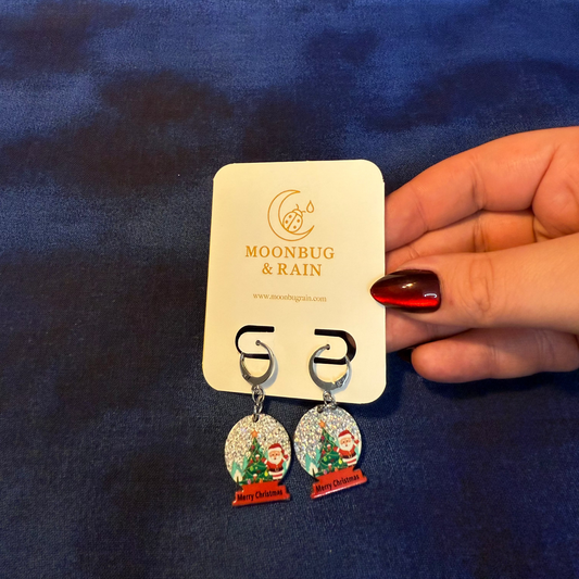 Earrings with snow globe design held by a hand against a blue background