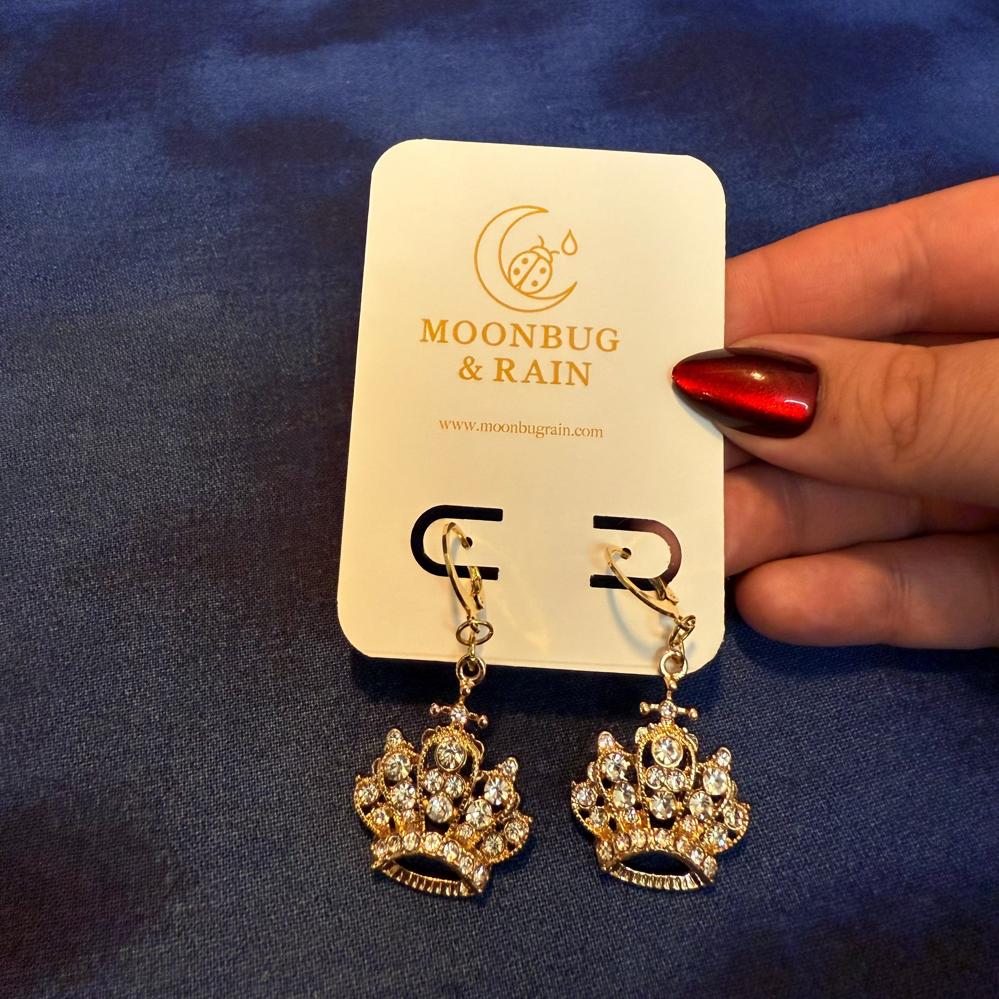 Gold earrings on a 'Moonbug & Rain' branded card against a blue background