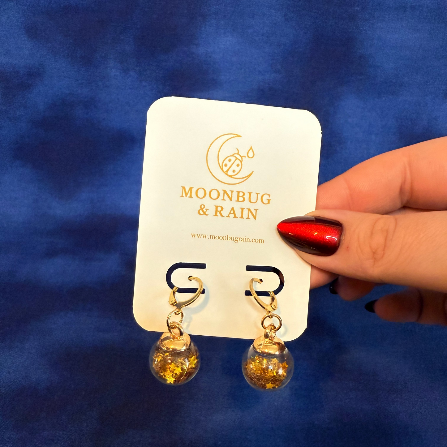 Gold earrings on a 'Moonbug & Rain' card against a blue background