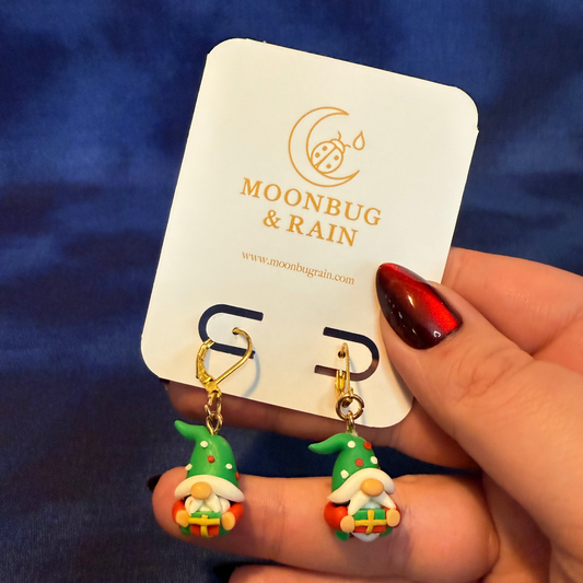 Pair of gnome-shaped earrings held by a hand with a blue background and 'Moonbug & Rain' card.