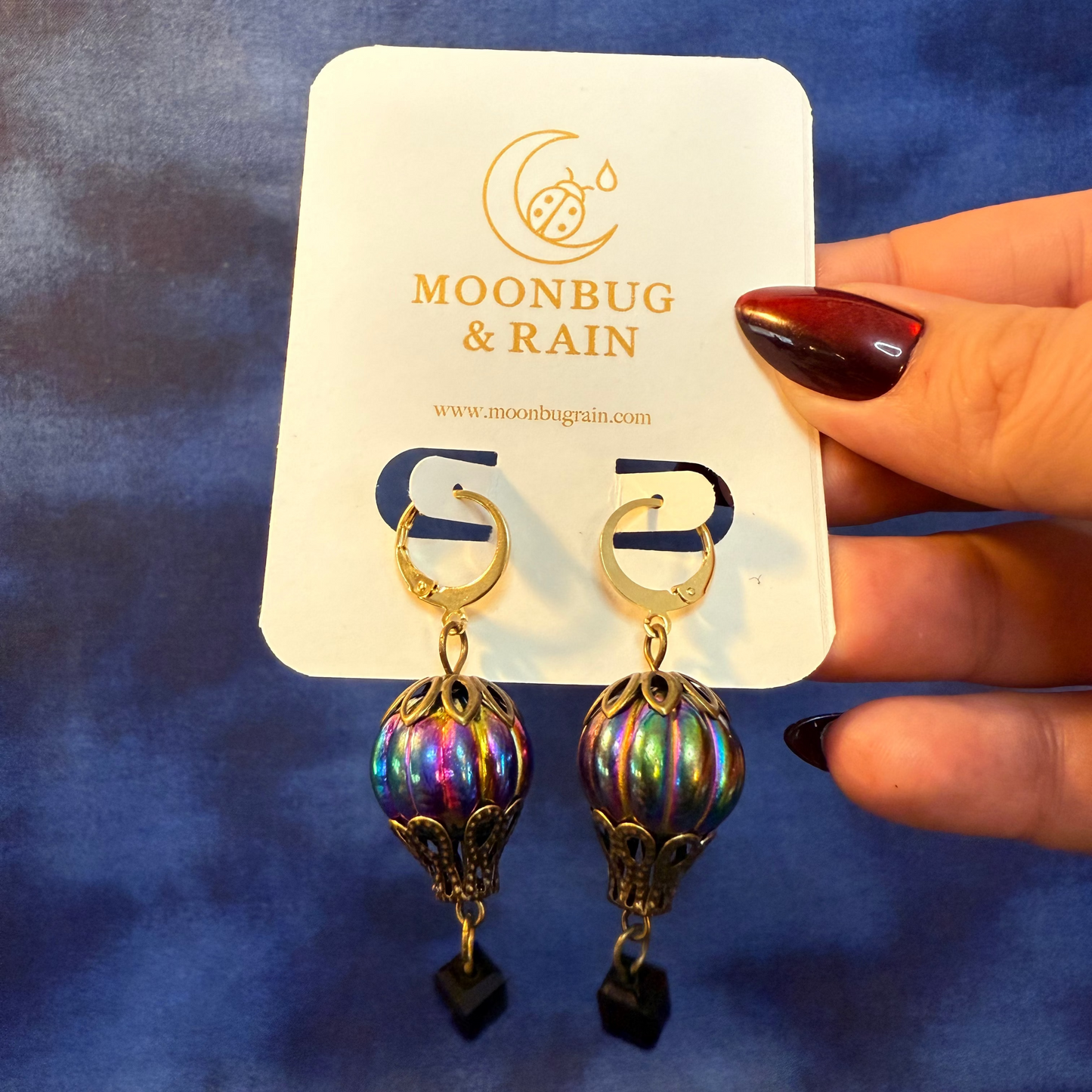 Multicolored earrings on a 'Moonbug & Rain' branded card against a blue background.
