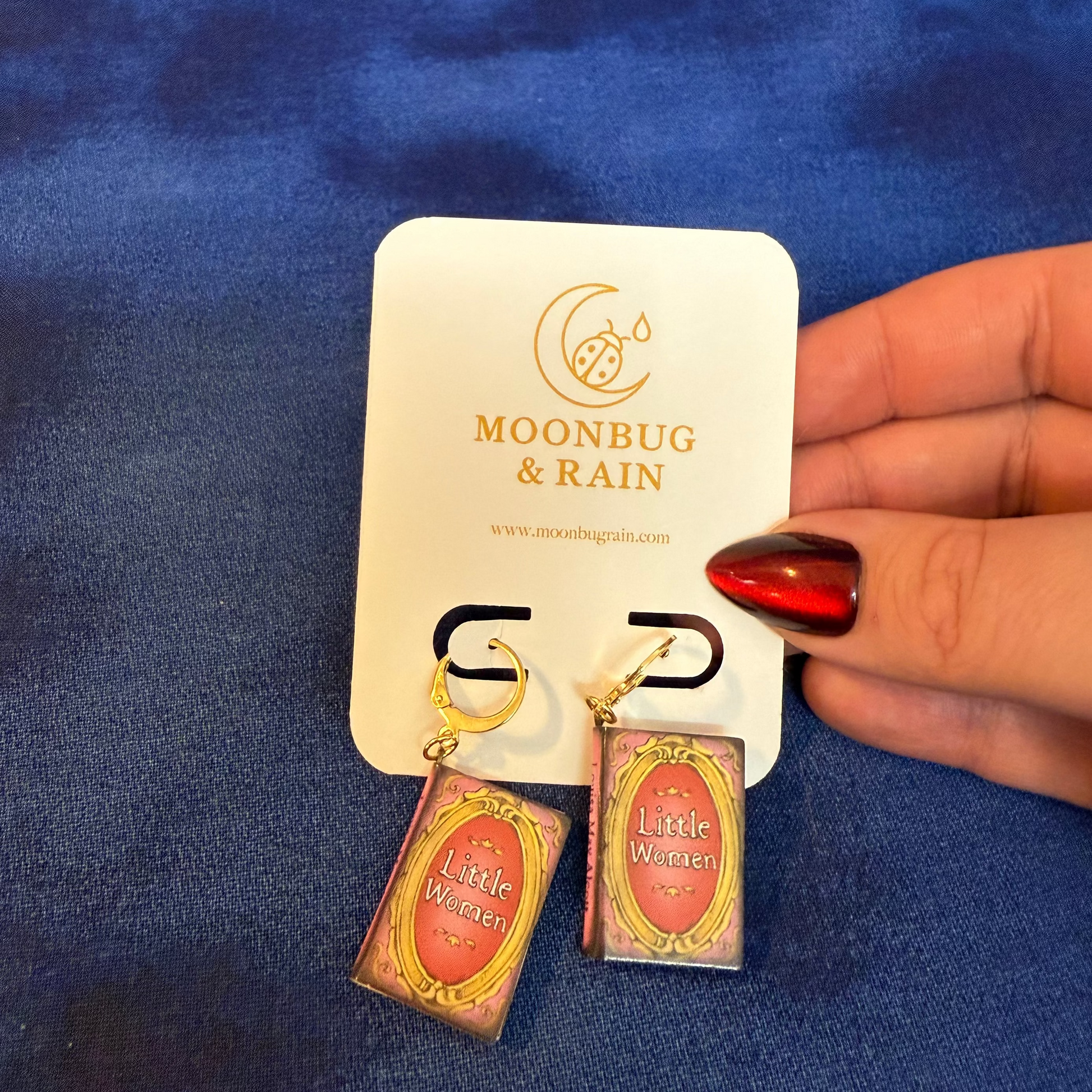 Earrings on a 'Moonbug & Rain' branded card held against a blue fabric background.