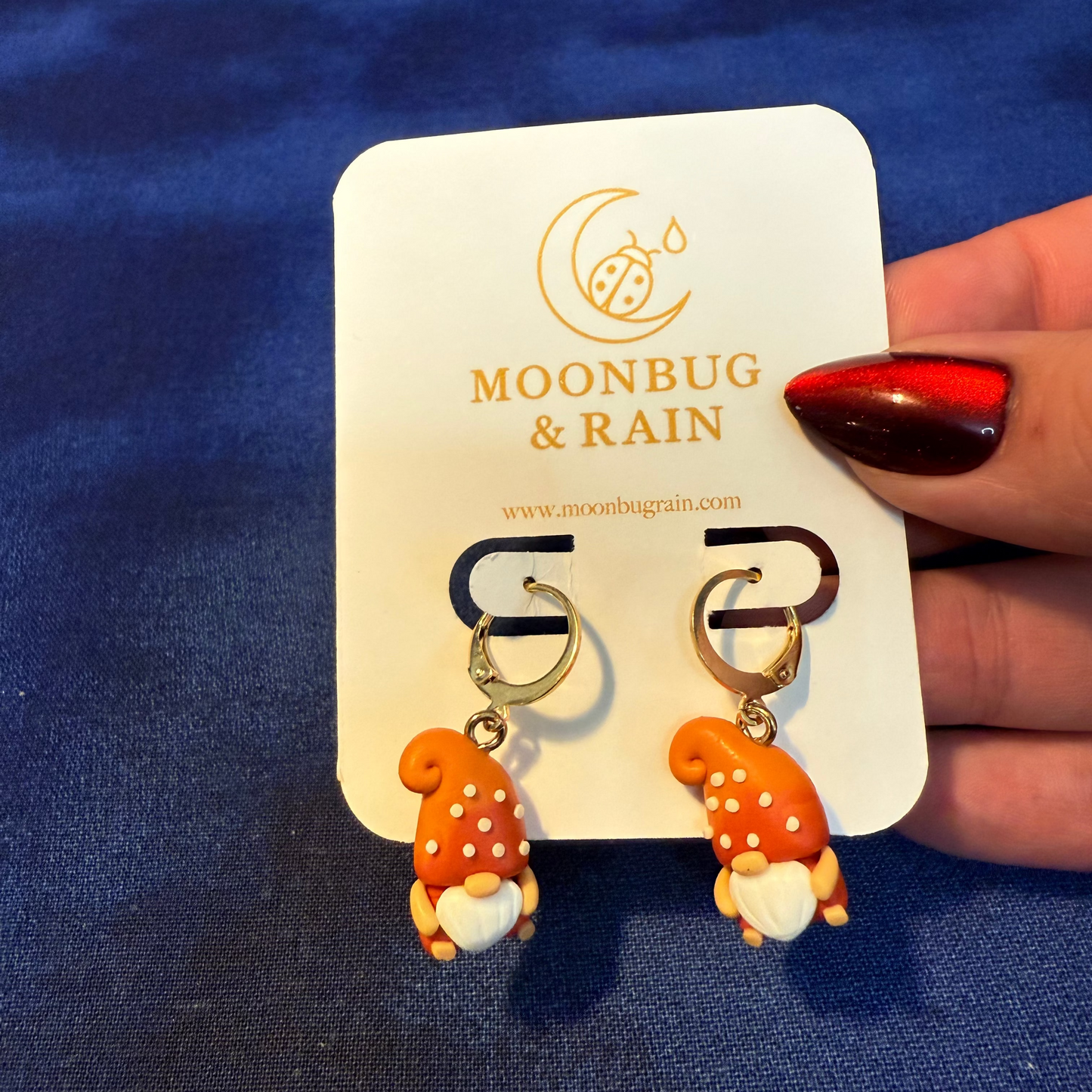 Pair of mushroom-shaped earrings on a Moonbug & Rain branded card against a blue background.