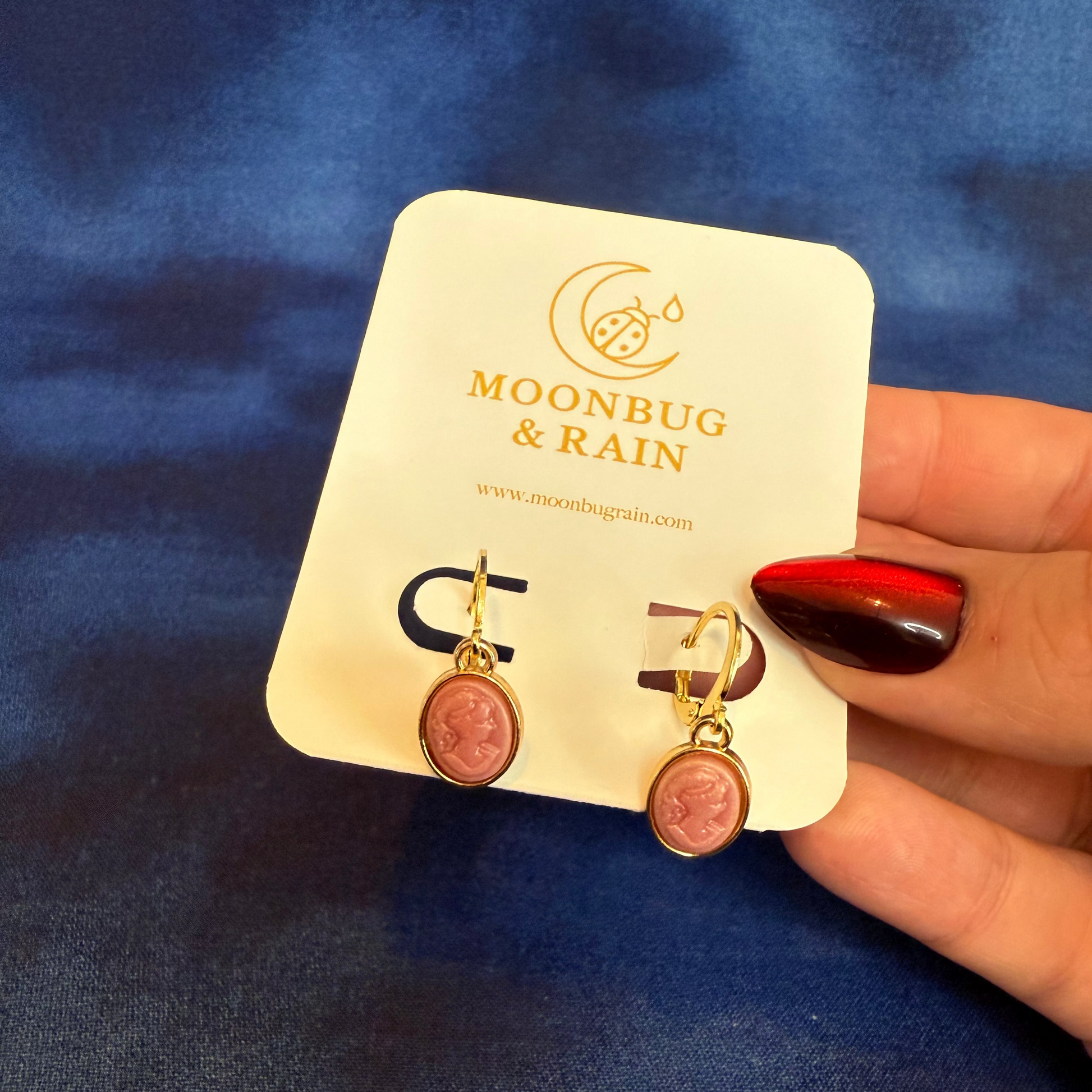 Pair of earrings on a 'Moonbug & Rain' branded card held by a hand against a blue fabric background.