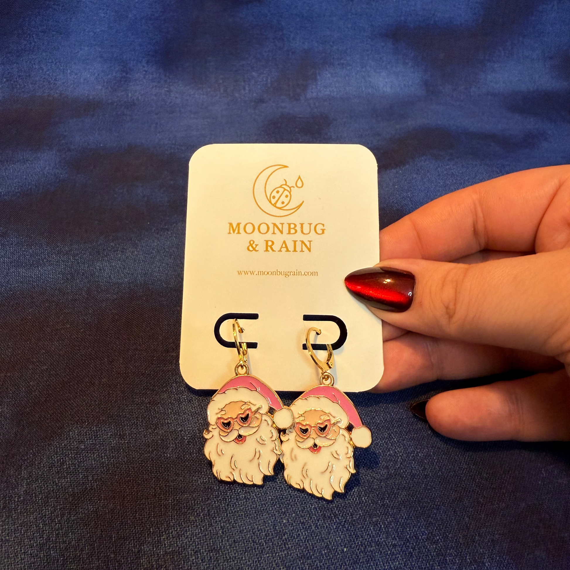 Santa Claus earrings on a 'Moonbug & Rain' card held by a hand against a blue fabric background