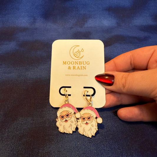 Santa Claus earrings on a 'Moonbug & Rain' card held by a hand against a blue fabric background
