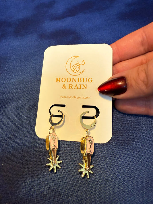 Gold earrings on a 'Moonbug & Rain' branded card against a blue fabric background.