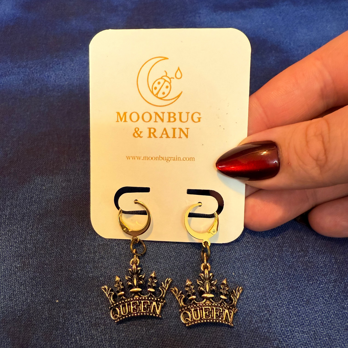 Pair of 'QUEEN' earrings on a Moonbug & Rain branded card held by a hand against a blue fabric background.