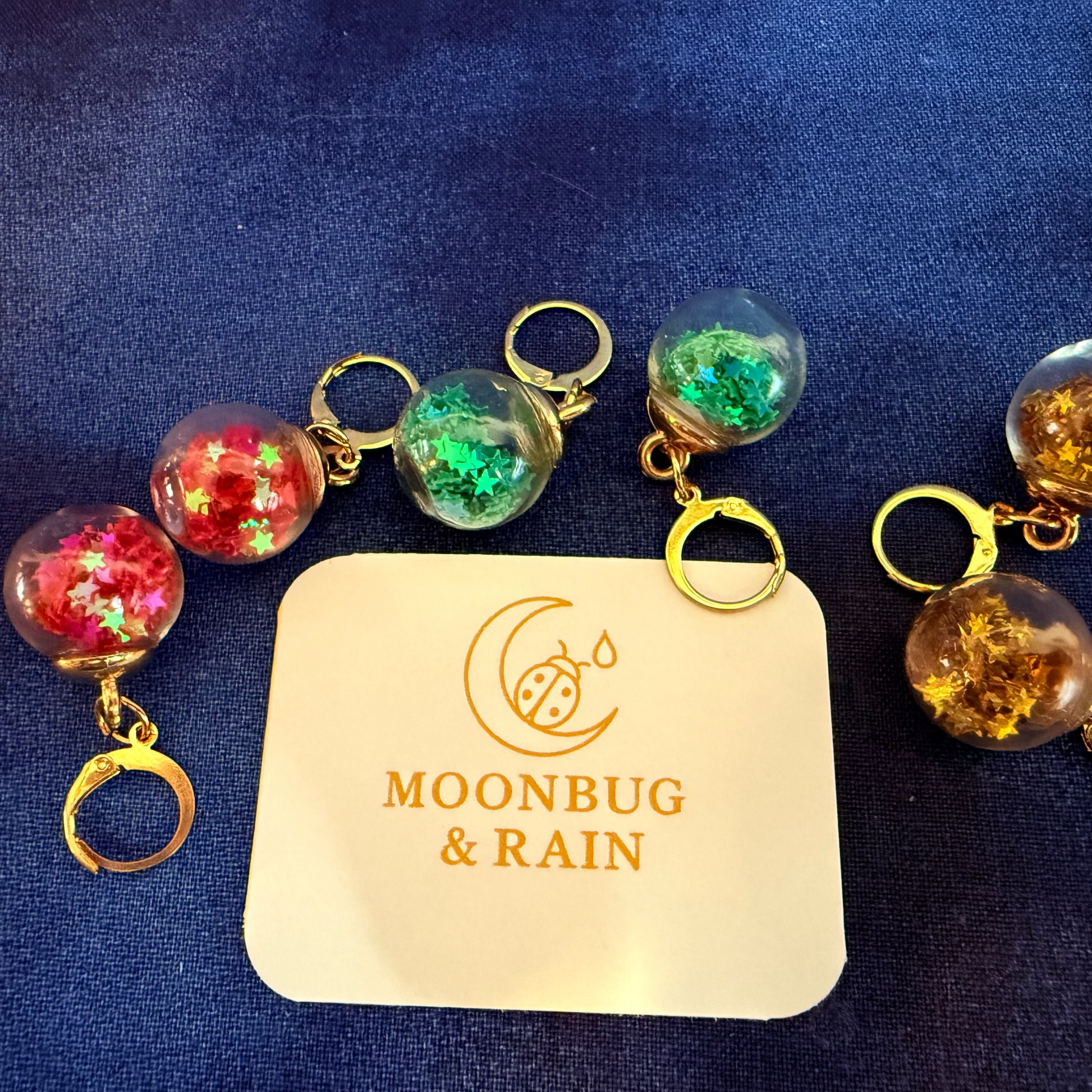 Decorative keychains with colorful beads on a blue fabric background, featuring the 'Moonbug & Rain' brand.