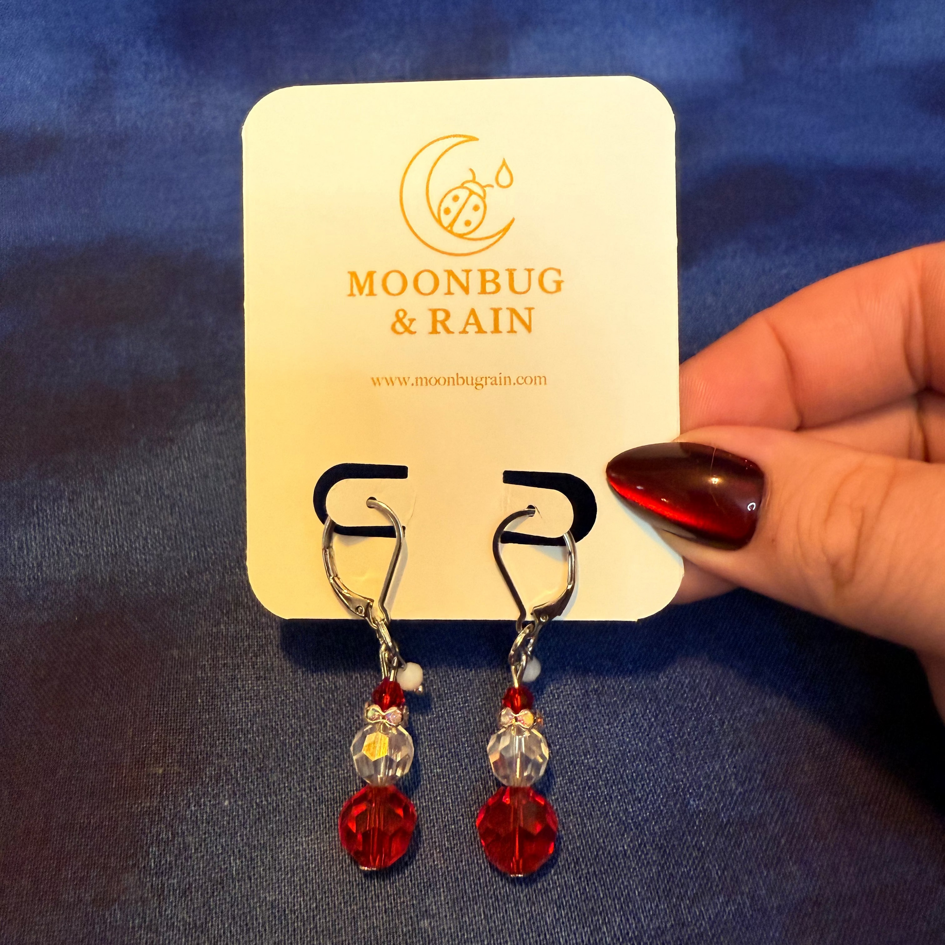 Red and clear crystal earrings on a 'Moonbug & Rain' branded card against a blue background.