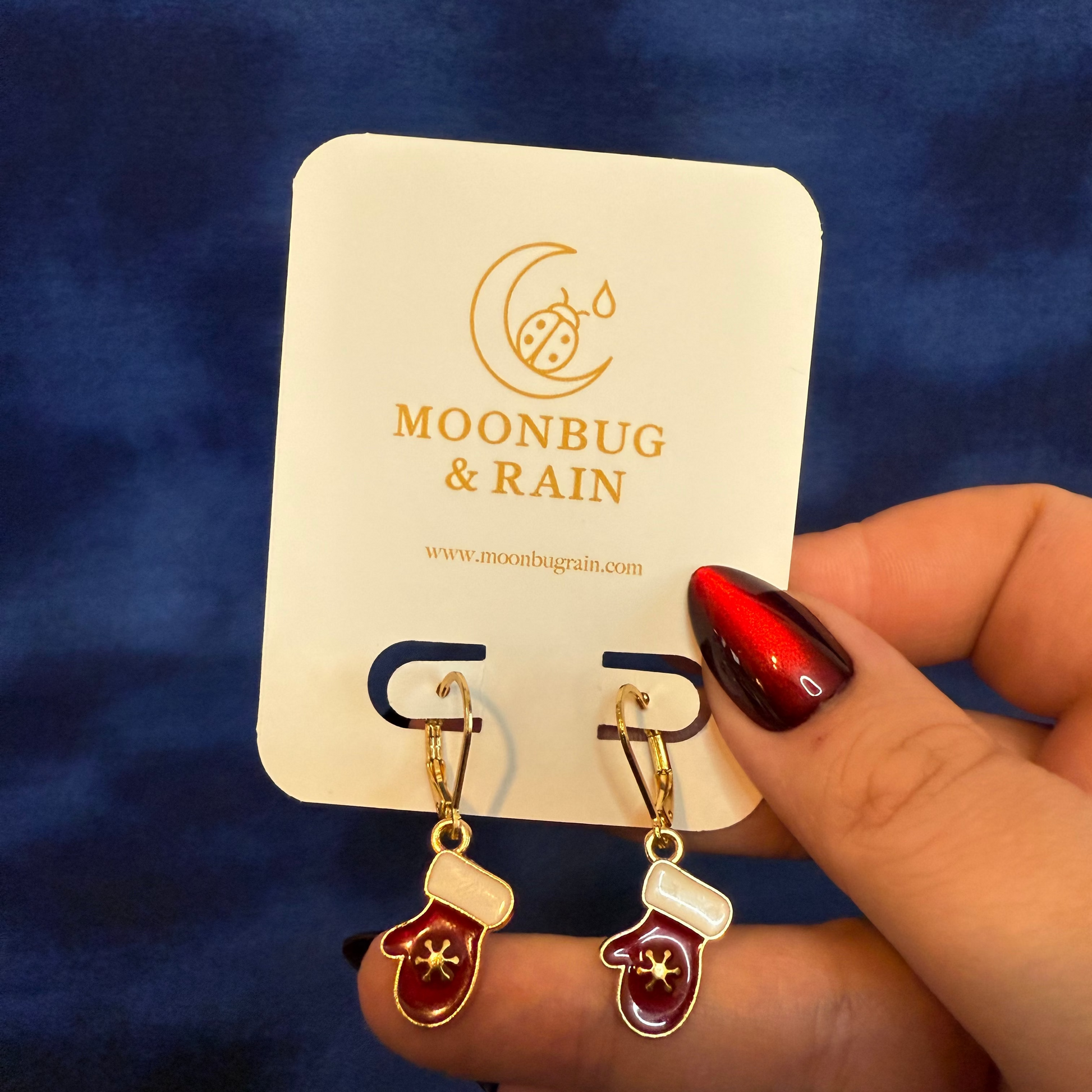 Earrings on a Moonbug & Rain branded card held by a hand with red nail polish against a blue background.