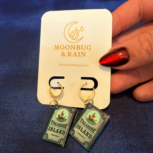 Earrings on a 'Moonbug & Rain' branded card held by a hand against a blue fabric background