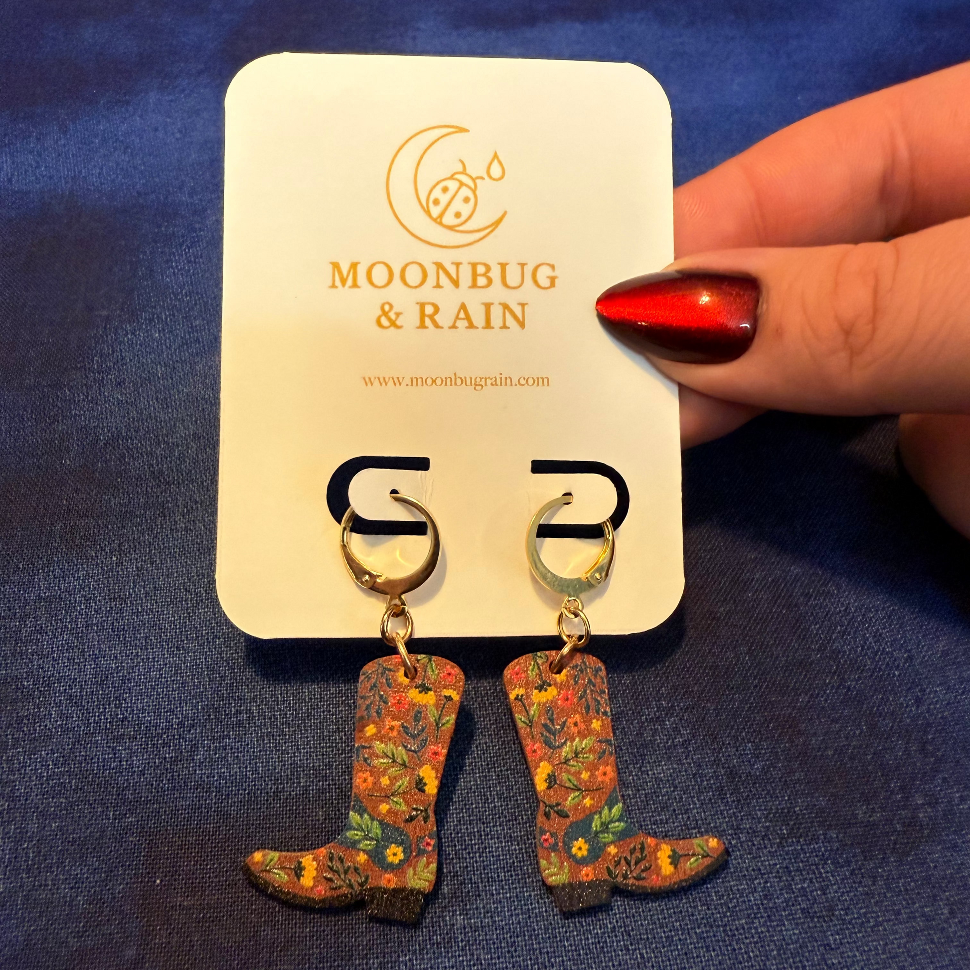 Cowboy boot earrings on a Moonbug & Rain branded card against a blue background.