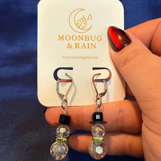 Pair of earrings held by a hand with a blue background, featuring the brand 'Moonbug & Rain'.