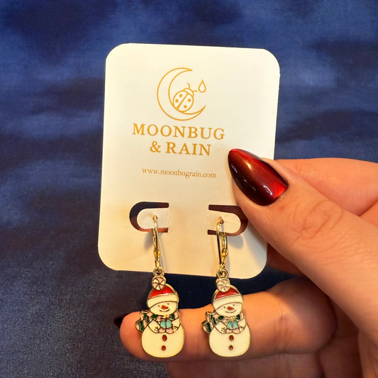 Snowman-themed earrings on a Moonbug & Rain card held by a hand with red nail polish against a blue background.