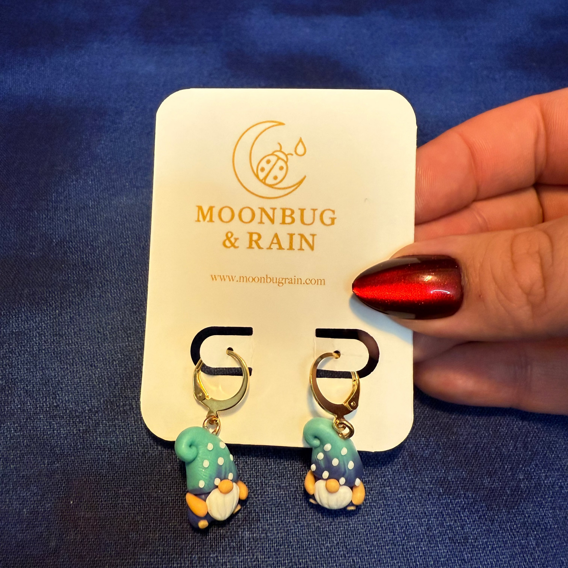 Pair of gnome-shaped earrings on a 'Moonbug & Rain' branded card held by a hand with red nail polish.