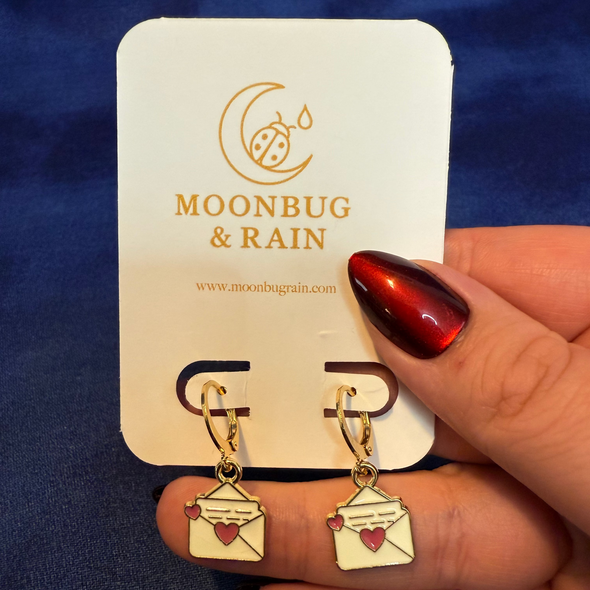 Gold earrings with heart designs held by a hand against a blue background, branded 'Moonbug & Rain'.