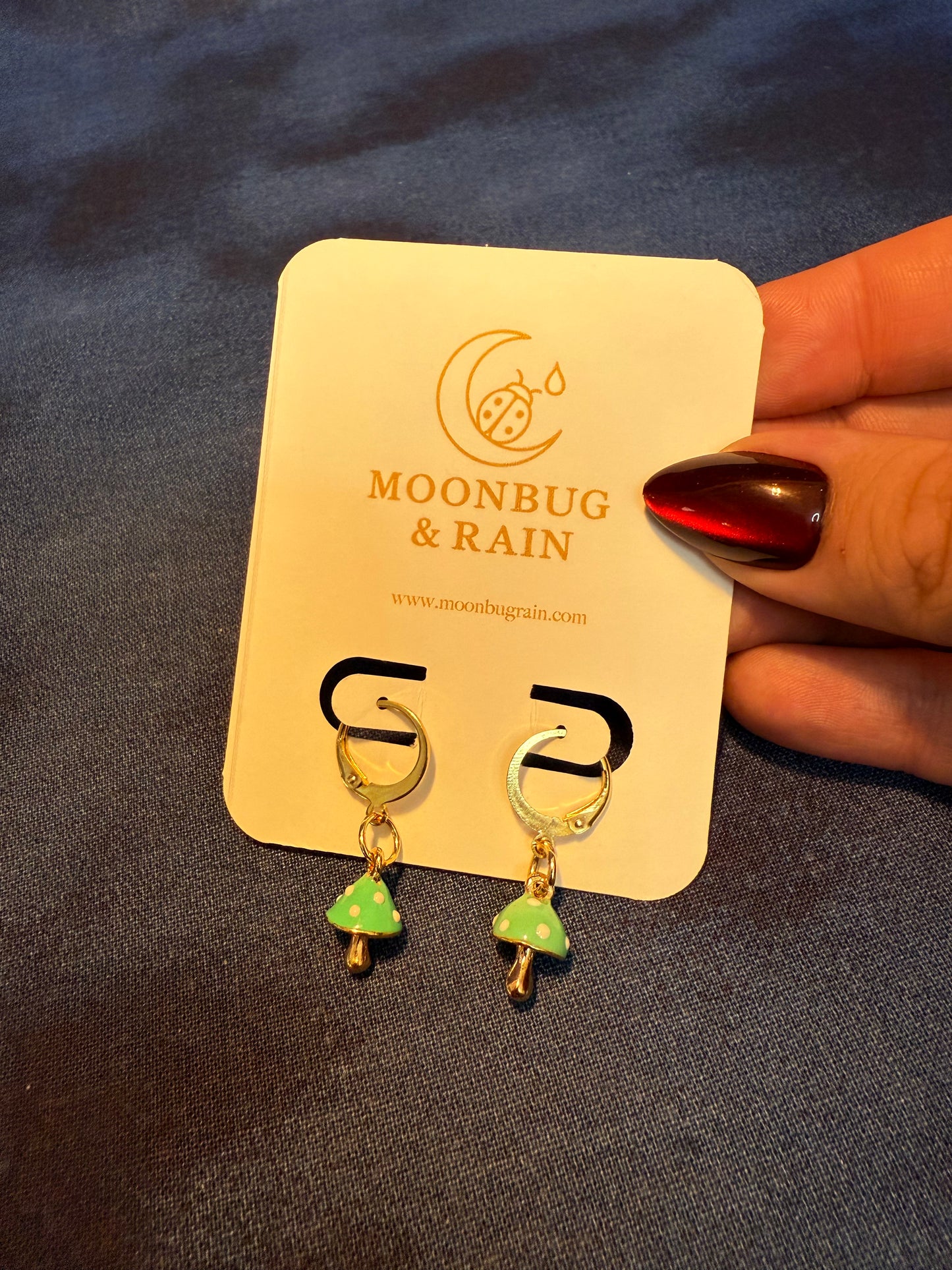 Gold and green earrings on a 'Moonbug & Rain' branded card held by a hand with red nail polish.