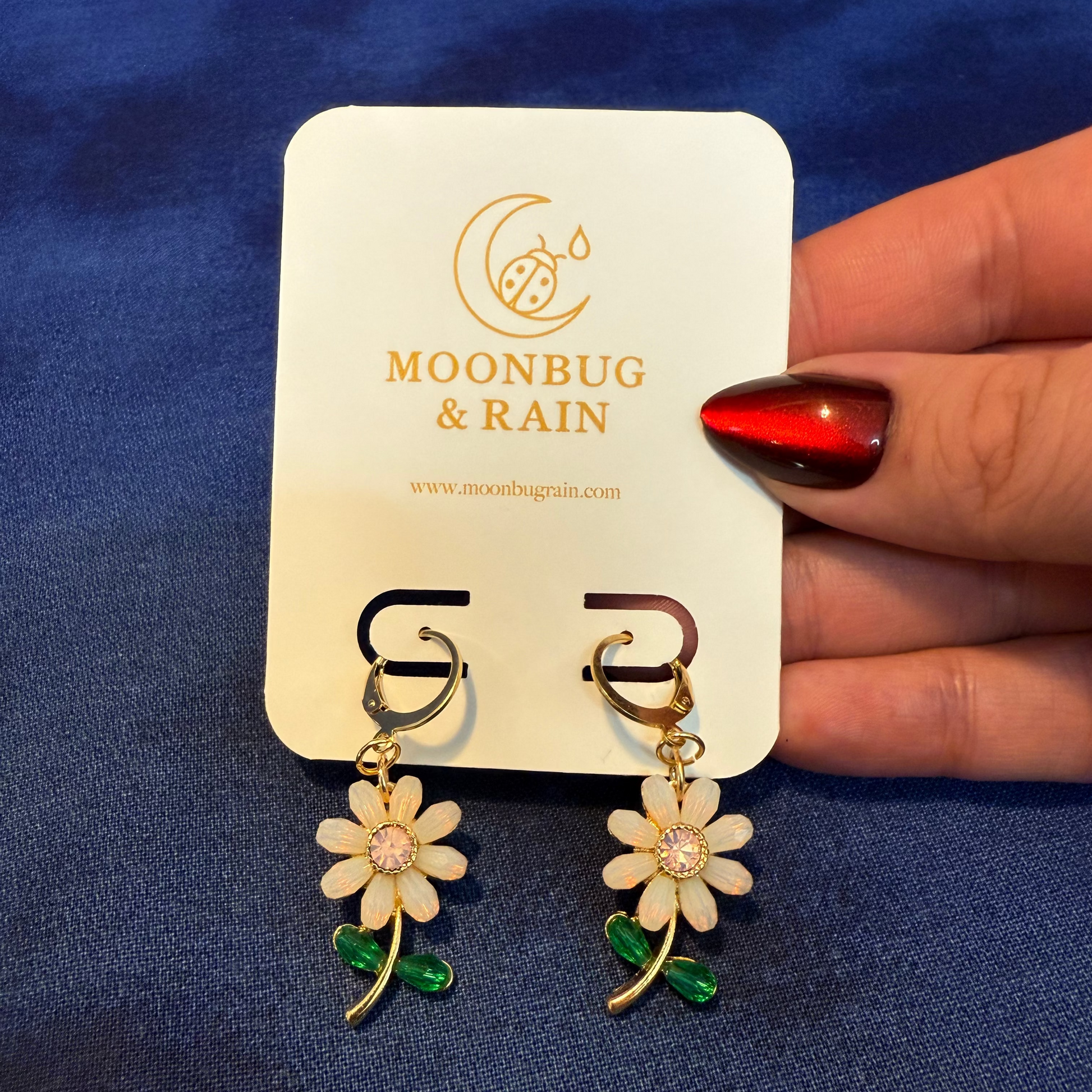 Flower-shaped earrings on a Moonbug & Rain branded card held by a hand with red nail polish against a blue background.
