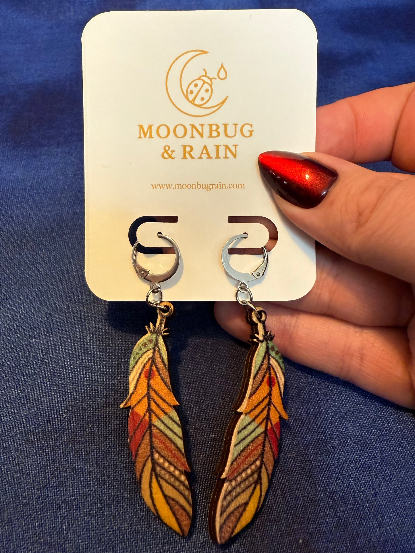 Feather-shaped earrings on a 'Moonbug & Rain' branded card held by a hand against a blue background.