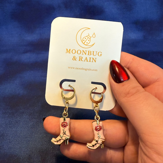 Gold earrings with red accents held by a hand against a blue background, with 'Moonbug & Rain' branding visible.
