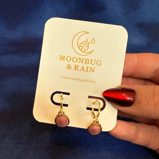 Earrings on a 'Moonbug & Rain' branded card held by a hand with red nail polish against a blue background.