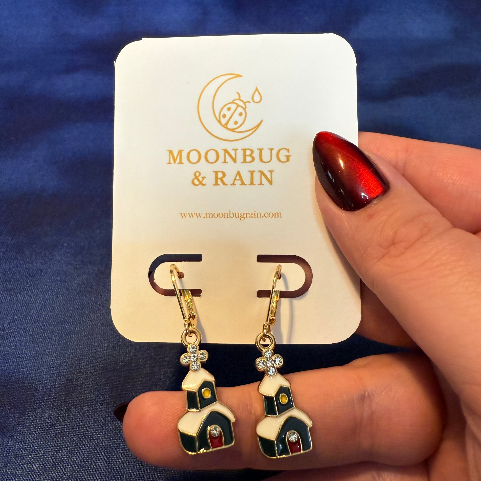 Earrings on a 'Moonbug & Rain' card held by a hand with red nail polish against a blue background.